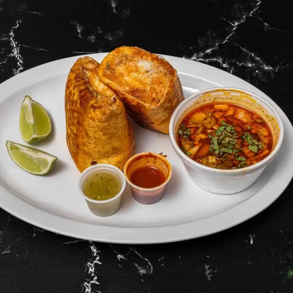 Two golden-brown burritos, a cup of red soup, two small sauce cups, and lime wedges on a white plate.