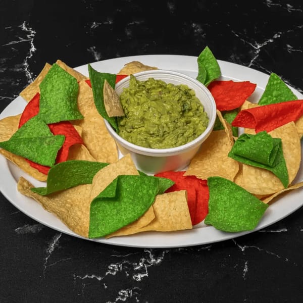A white plate of tri-color tortilla chips with a cup of guacamole on a black marble surface.