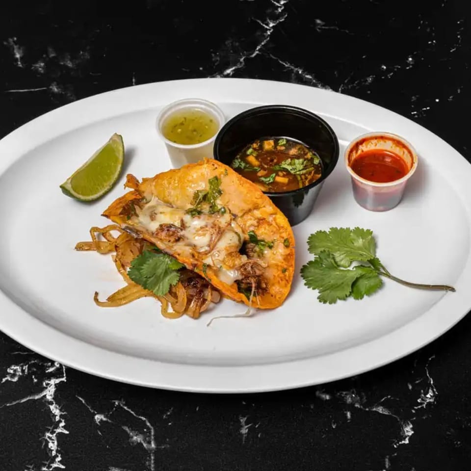 A Tacos El Primo birria taco with melted cheese, onions, cilantro, lime, and two dipping sauces.