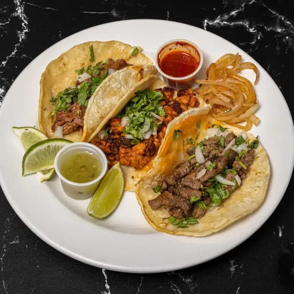 A plate of three Tacos El Primo tacos with various meats, cilantro, onions, lime, and sauces.