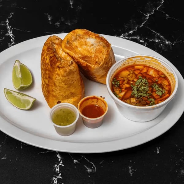 Two golden-brown burritos, a cup of soup, two sauces, and lime wedges on a white plate.